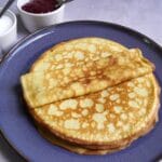Stak gyldne, klassiske tynde pandekager foldet på en blå tallerken, serveret med sukker og hindbærmarmelade ved siden af
