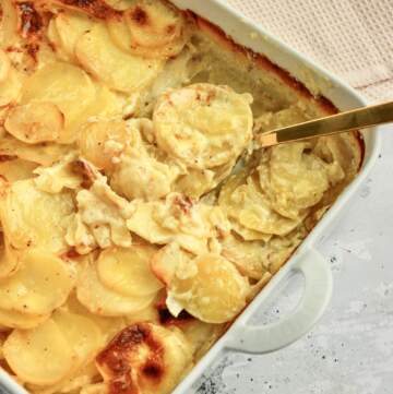 Cremede flødekartofler i ovn i et hvidt ovnfast fad, hvor en guld ske løfter en portion med bløde kartoffelskiver og cremet flødesauce