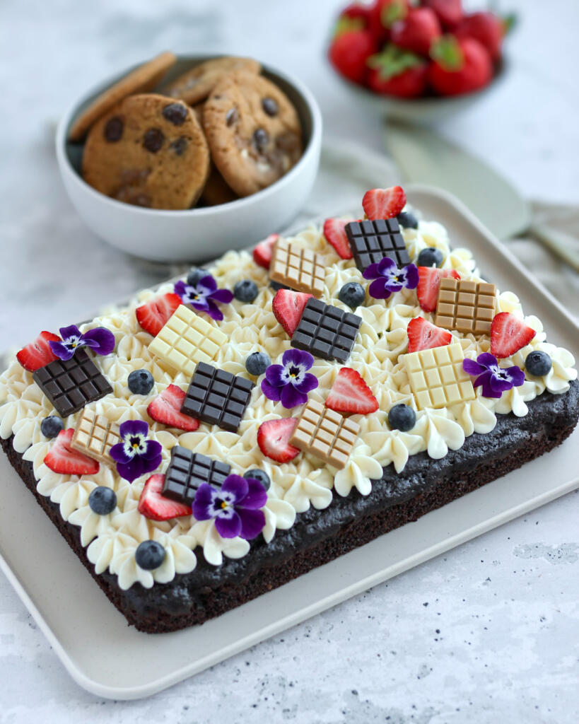 Chokoladekage med cream cheese frosting toppet med jordbær, blåbær, chokoladebarer og spiselige blomster serveret på lyst fad med chokoladekiks i baggrunden
