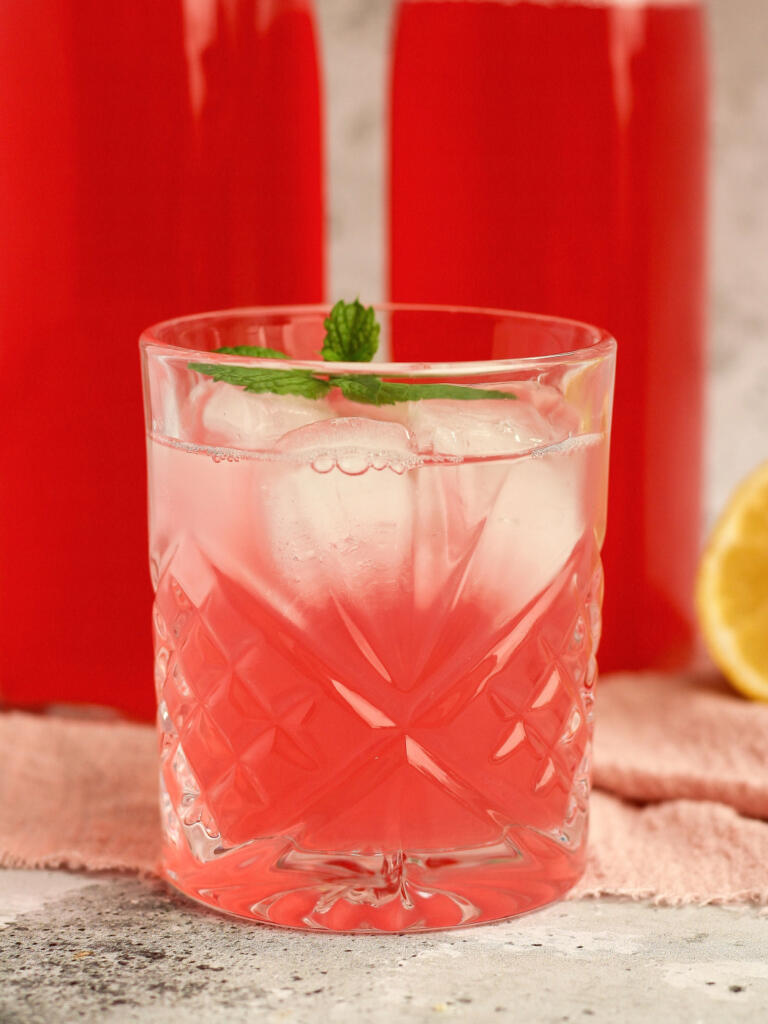Glas med kold rabarbersaft blandet med vand og isterninger foran flasker med koncentreret hjemmelavet rabarbersaft