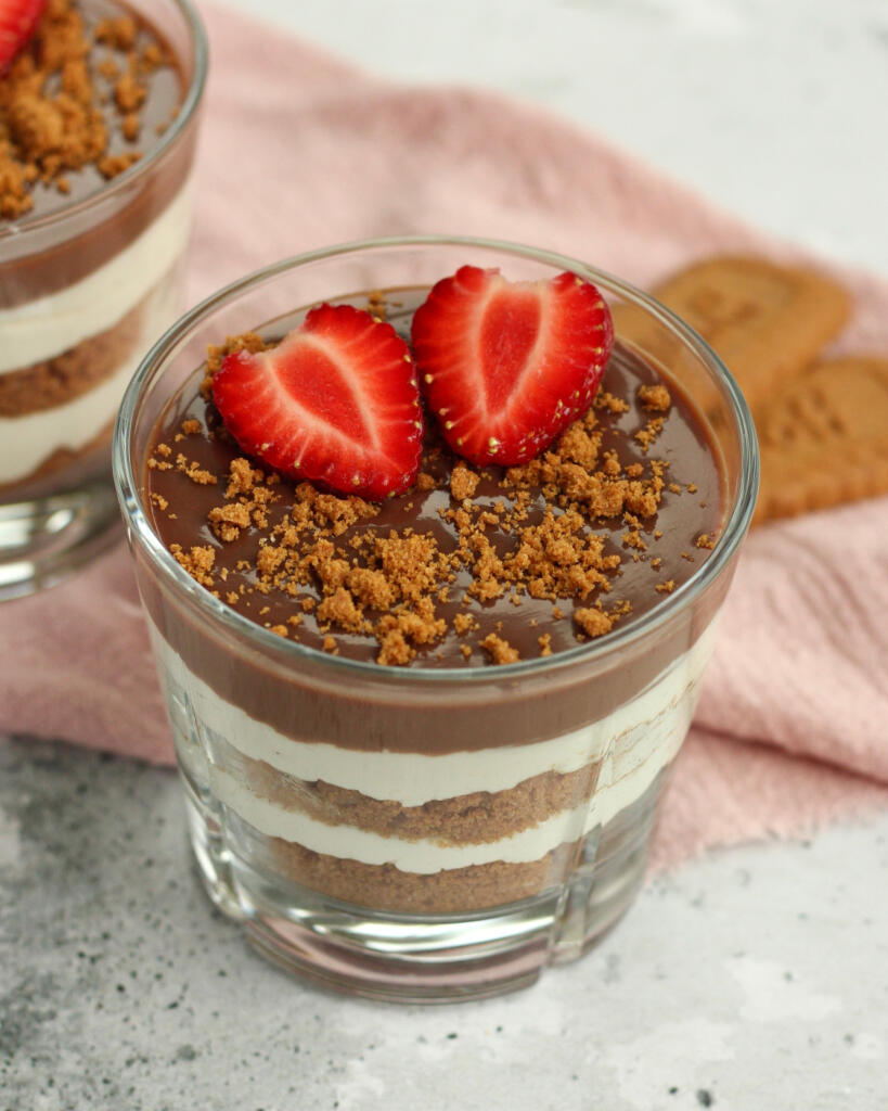 Biscoff cheesecake i glas med synlige lag af knuste Biscoff kiks, cheesecakecreme og chokoladeganache pyntet med jordbær