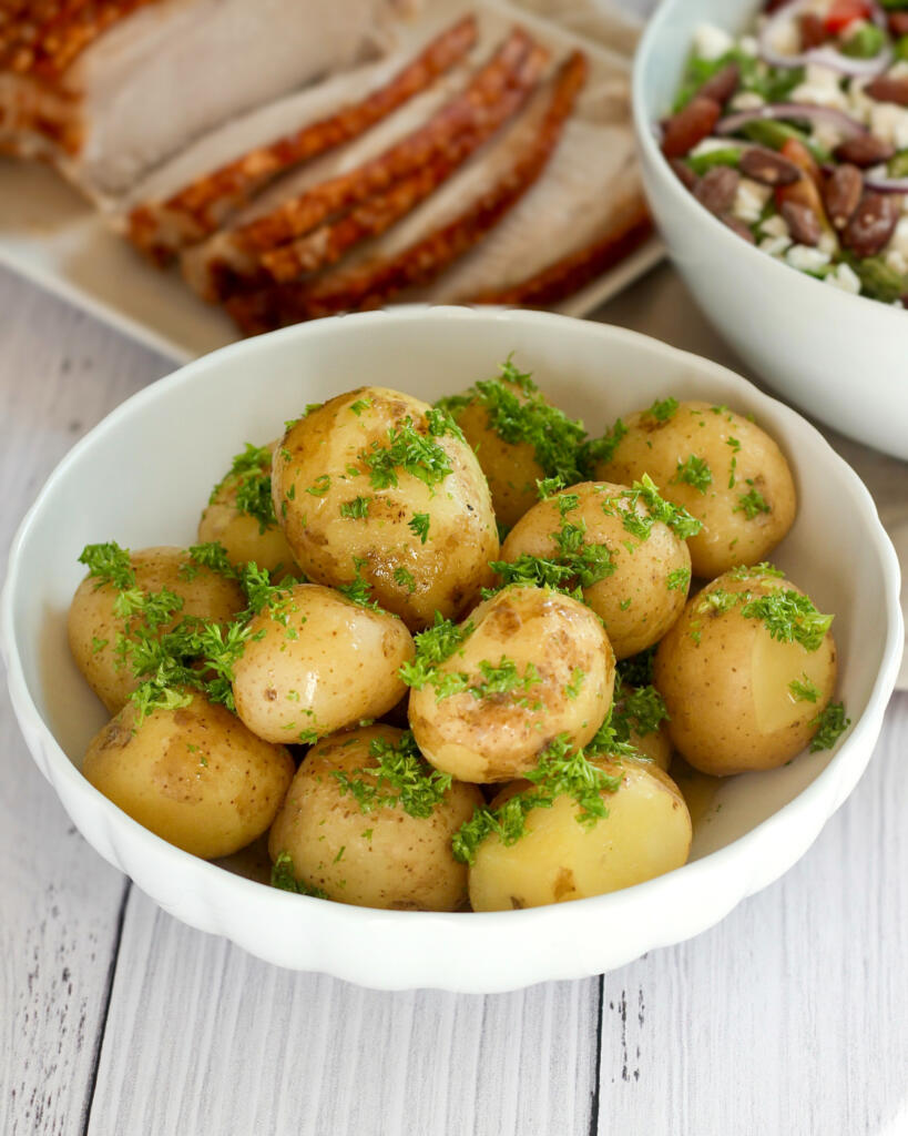 Nye kartofler med persille og smeltet smør serveret i skål med flæskesteg og salat i baggrunden