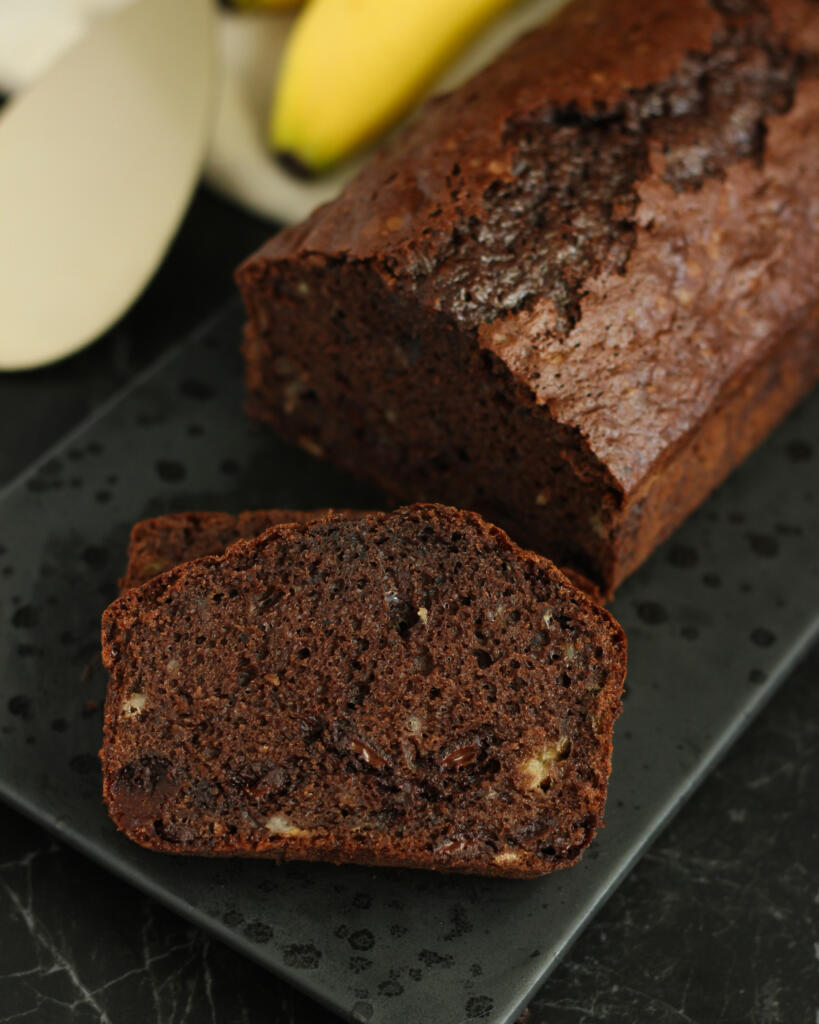 Svampet chokoladekage med banan i brødform skåret i skiver med friske bananer i baggrunden